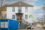 Vor einem zweigeschossigen weißen Gebäude mit zentralem Eingang und Außentreppe steht links ein großer blauer Container, während rechts an der Fassade ein buntes dreieckiges Logo angebracht ist.