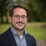 Ein dunkelhaariger Mann mit Brille im Portrait während er im Freien steht.