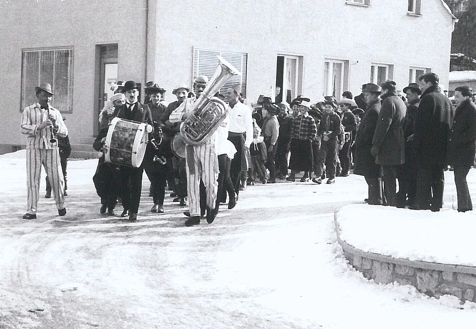 Das historische Schwarzweißfoto zeigt einen Faschingszug im Jahr 1958, bei dem eine Musikgruppe in gestreiften Kostümen voran marschiert, gefolgt von zahlreichen Zuschauern und Teilnehmern entlang einer verschneiten Straße.
