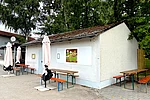 Ein kleines Café mit heller Fassade und Sitzplätzen im Außenbereich liegt an einem Platz mit Bäumen und lädt mit Tischen und Stühlen zum Verweilen ein.