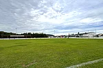 Ein großer Fußballplatz mit grünem Rasen und weißen Toren liegt neben einem Vereinsheim und Zuschauerbereich, während im Hintergrund Bäume und weitere Gebäude der Sportanlage zu sehen sind.
