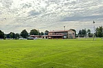 Ein gepflegter Fußballplatz mit grünem Rasen und weißem Tor liegt vor einer Baumreihe am Rand des Sportgeländes, während im Hintergrund Werbebanden und weitere Bereiche der Anlage zu sehen sind.