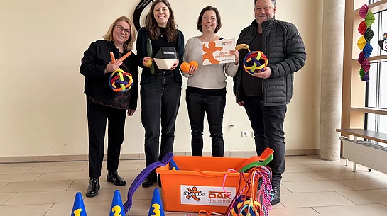 Vier Personen stehen in einem Innenraum nebeneinander, lächeln in die Kamera und präsentieren bunte Spiel- und Bewegungsmaterialien wie Bälle, Seile und Markierungshütchen aus einer großen orangefarbenen Kiste.