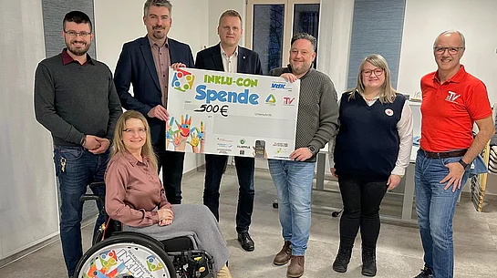 Eine Gruppe von sieben Personen präsentiert gemeinsam einen großen Spendenscheck über 500 Euro, wobei eine Frau im Rollstuhl vorne sitzt und alle freundlich in die Kamera lächeln.