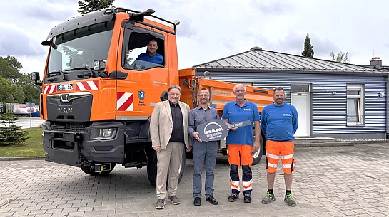 Mehrere Personen stehen vor einem neuen orangefarbenen MAN-Kommunalfahrzeug des städtischen Bauhofs. Ein Mitarbeiter sitzt im Führerhaus, während Vertreter der Stadt, der Herstellerfirma und Bauhofmitarbeiter vor dem Fahrzeug für ein Übergabefoto posieren. Der neue LKW ist auf dem Gelände des Bauhofs abgestellt.