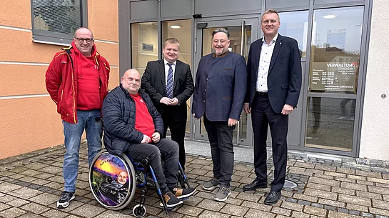 Vor dem Eingang eines Verwaltungsgebäudes stehen mehrere Männer zusammen, darunter eine Person im Rollstuhl, und posieren lächelnd für ein gemeinsames Gruppenfoto.