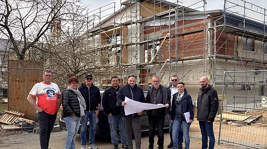 Mehrere Personen stehen vor einem Gebäude im Bau mit Gerüst und Baukran und betrachten gemeinsam einen ausgebreiteten Bauplan.