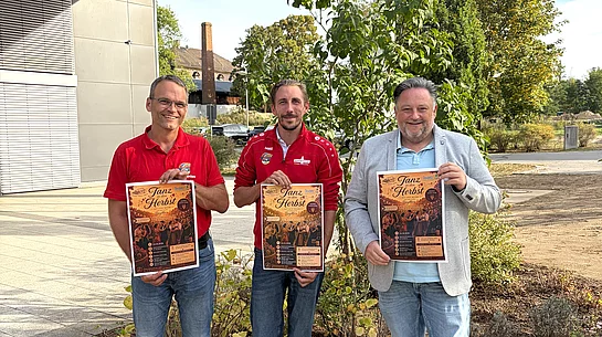 Zwei Vertreter der FG D’Rummlfelser und Bürgermeister Thomas Beer stehen vor der Dreifachturnhalle und halten jeweils ein Plakat für die Veranstaltung „Tanz in den Herbst“ in den Händen.