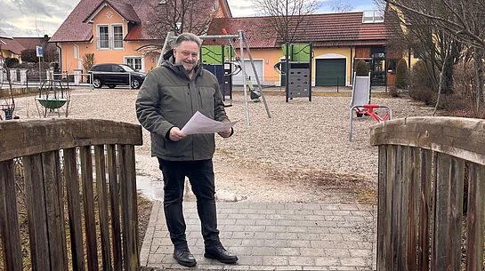 Ein Mann steht auf einer kleinen Holzbrücke vor einem Spielplatz und hält ein Blatt Papier in der Hand, während hinter ihm Spielgeräte und Wohnhäuser zu sehen sind.