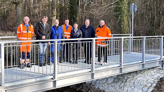 Eine Gruppe von Personen steht gemeinsam auf einer neu errichteten kleinen Brücke über einen Bach vor einem bewaldeten Hintergrund.