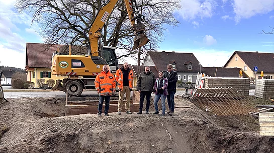 Vor einem großen Bagger stehen mehrere Personen am Rand einer ausgehobenen Baugrube, in der ein Brückenbau vorbereitet wird.