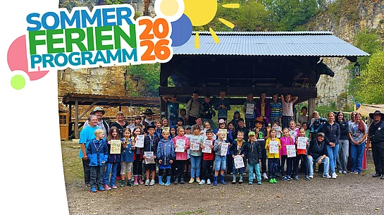 Eine große Gruppe von Kindern und Erwachsenen steht bei einem Kinder-Rodeo im Freien vor einer überdachten Holzhütte; viele Kinder halten Urkunden in den Händen.