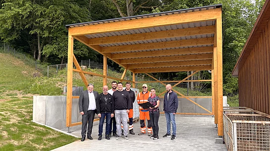 Eine Gruppe von acht Personen steht unter einem neu errichteten, offenen Carport aus Holz mit Metalldach auf einem gepflasterten Platz. Einige tragen Alltagskleidung, zwei Personen auffällige orangefarbene Arbeitskleidung. Im Hintergrund befinden sich ein bewaldeter Hang sowie technische Anlagen. Rechts sind große Metallcontainer zu sehen. Die Gruppe blickt in die Kamera und steht mittig unter der Konstruktion, die den Eindruck einer offiziellen Besichtigung oder Fertigstellung am Wasserwerk vermittelt.