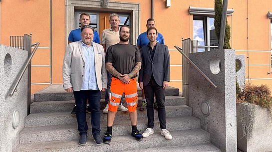 Sechs Männer stehen für ein Gruppenfoto auf der Treppe vor einem orangefarbenen Gebäude. Im Mittelpunkt steht der neue Bauhofmitarbeiter Patrick Mailbeck in auffälliger orangefarbener Arbeitshose. Neben und hinter ihm stehen fünf Männer, die ihn zum Beginn seiner neuen Tätigkeit willkommen heißen.