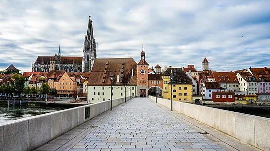 Eine gepflasterte Brücke führt auf eine historische Altstadt mit bunten Häusern, einem Stadttor und dem Dom im Hintergrund unter bewölktem Himmel zu.