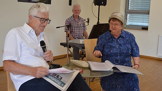 In einem Veranstaltungsraum sitzen ein Mann mit Mikrofon und Unterlagen sowie eine Frau mit geöffnetem Ordner an einem kleinen Tisch, während im Hintergrund ein weiterer Mann an einem Keyboard sitzt und die Szene musikalisch begleitet.