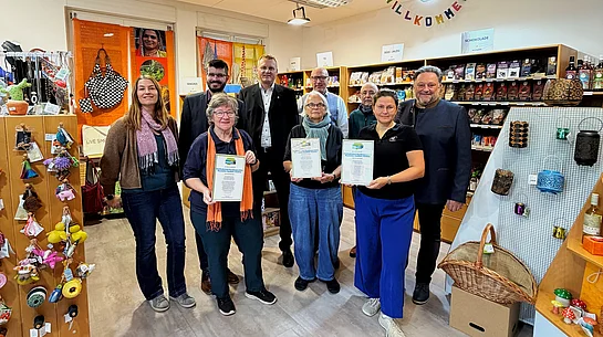 Gruppenfoto im Weltladen Teublitz anlässlich der Verlängerung der Fairtrade-Auszeichnung für das Städtedreieck bis 2028. Neun Personen, fünf Männer und vier Frauen, stehen in zwei Reihen vor Verkaufsregalen mit fair gehandelten Lebensmitteln und Produkten. Drei Frauen in der vorderen Reihe halten gerahmte Urkunden zur erneuten Auszeichnung in den Händen. Im Hintergrund sind Regale mit Schokolade, Tee und weiteren Waren sowie eine bunte Willkommensgirlande zu sehen. Die Gruppe blickt in die Kamera.