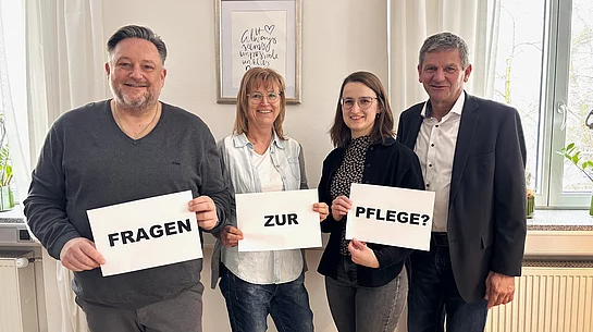 Vier Personen stehen nebeneinander in einem hellen Raum und halten jeweils ein Blatt Papier hoch, sodass zusammen die Frage „Fragen zur Pflege?“ entsteht, während sie freundlich in die Kamera blicken.