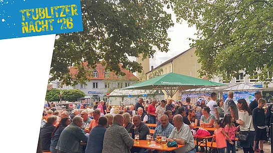 Grafik zur „Teublitzer Nacht ’26“ mit einer stimmungsvollen Aufnahme des Marktplatzes in Teublitz. Zahlreiche Besucherinnen und Besucher sitzen und stehen an Biertischen unter großen Bäumen und Sonnenschirmen, unterhalten sich und genießen den Abend. Im Hintergrund sind Marktstände und Gebäude rund um den Platz zu sehen. Oben links steht auf blauem Hintergrund der Schriftzug „Teublitzer Nacht ’26“.