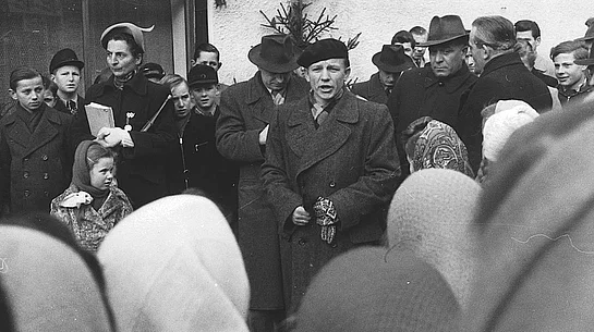 Eine historische Schwarz-Weiß-Aufnahme zeigt einen Redner mit Baskenmütze, der während der Eröffnungsfeier vor einer großen Menschenmenge spricht. Um ihn herum stehen Erwachsene und Jugendliche, während zahlreiche Besucherinnen und Besucher aufmerksam zuhören.