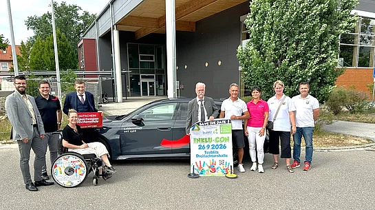 Vor der Dreifachsporthalle in Teublitz steht eine Gruppe von neun Personen neben einem dunklen Elektrofahrzeug. Eine Person sitzt im Rollstuhl. Mehrere Beteiligte halten ein Veranstaltungsplakat mit der Aufschrift „Inklu-Con 2026“ sowie einen Gutschein für einen Sportwagen. Die Gruppe lächelt in die Kamera. Das Foto entstand bei der offiziellen Vorstellung der Inklu-Con 2026, einer inklusiven Veranstaltung im Städtedreieck.