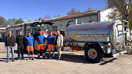 Mehrere Männer stehen bei Sonnenschein vor einem Bauhof und präsentieren gemeinsam einen Traktor mit Gießanhänger.
