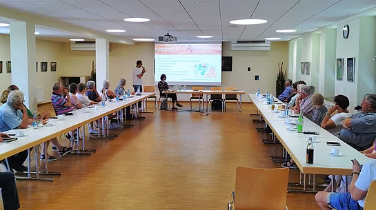 Mehrere ältere Menschen sitzen an Tischen in U-Form in einem hellen Raum und hören einem Vortrag zu, der mit einer Präsentation auf einer Leinwand unterstützt wird.