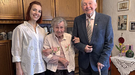 Eine jüngere Frau steht lächelnd neben einem älteren Ehepaar in einem Wohnzimmer, während die Frau den Arm der Seniorin hält und der Mann einen Gehstock in der Hand hat.