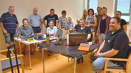 Eine Gruppe von rund einem Dutzend Personen steht und sitzt in einem hellen Raum um mehrere Tische, auf denen verschiedene Elektrogeräte, Werkzeugkästen, Messgeräte und Kleinteile liegen. Im Mittelpunkt steht ein geöffnetes Hi-Fi-Gerät, an dem gearbeitet wird. Die Teilnehmenden lächeln in die Kamera und vermitteln den Eindruck einer gemeinschaftlichen und engagierten Atmosphäre. Das Foto entstand anlässlich des ersten Geburtstags des Reparatur-Cafés.