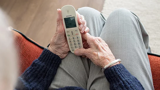 Eine ältere Person sitzt in einem Sessel und hält ein schnurloses Telefon in beiden Händen. Das Symbolbild begleitet die Ankündigung eines Vortrags zum Thema Schockanrufe und Telefonbetrug.