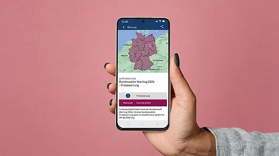 Symbolbild zum Bundesweiten Warntag 2026: Eine Hand hält ein Smartphone vor rosafarbenem Hintergrund, auf dessen Display eine Karte Deutschlands und eine Probewarnung zum Warntag am 10. September 2026 zu sehen sind.