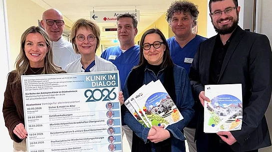 Mehrere Personen stehen gemeinsam auf einem Krankenhausflur, lächeln in die Kamera und präsentieren ein großes Veranstaltungsplakat sowie Programmhefte zu einer medizinischen Vortragsreihe.