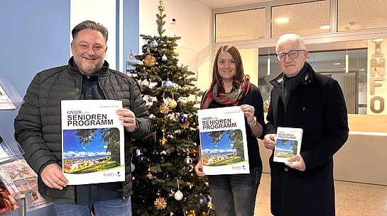 Drei Personen stehen in einem Eingangsbereich vor einem geschmückten Weihnachtsbaum und halten jeweils das neue Seniorenprogramm in der Hand. Die Hefte zeigen ein Titelbild mit Landschaftsfotografie. Alle drei lächeln in die Kamera, und im Hintergrund ist ein Infobereich des Gebäudes zu sehen.