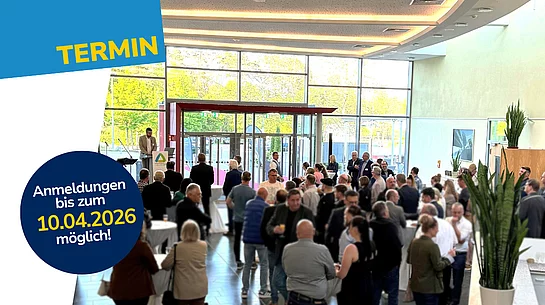 In einer großen, hellen Eingangshalle mit Glasfront stehen zahlreiche Menschen an Stehtischen und unterhalten sich, während links ein Redner an einem Pult spricht und darüber ein grafischer Hinweis mit Anmeldedatum eingeblendet ist.