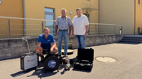 Ein Mitarbeiter des Klärwerks kniet vor einer neu angeschafften Kanal-Kamera mit Fahrwagensystem und Kontrolltechnik, während zwei Vertreter des Zweckverbands hinter ihm stehen. Die drei begutachten gemeinsam die moderne Ausrüstung, die künftig für die Inspektion von Abwasserkanälen eingesetzt wird.