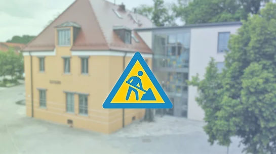 Im Vordergrund befindet sich ein großes, dunkelblaues Baustellensymbol – ein Dreieck mit einer stilisierten Person, die mit einer Schaufel arbeitet. Der Hintergrund ist weichgezeichnet und zeigt das Rathaus von Teublitz in warmen Gelb- und Beigetönen.