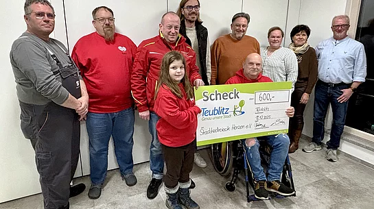 Auf dem Foto steht eine Gruppe von Menschen zusammen, während ein Mädchen und ein Mann im Rollstuhl symbolisch einen großen Spendenscheck über 600 Euro präsentieren.