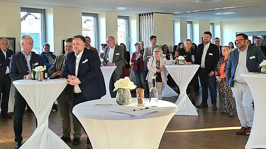 Eine Gruppe von Menschen steht bei einem Empfang in einem hellen Raum an Stehtischen zusammen und hört aufmerksam zu.