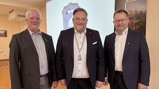 Drei Männer stehen nebeneinander und blicken in die Kamera. Der Mann in der Mitte trägt eine Amtskette und steht damit erkennbar im Mittelpunkt. Alle drei sind formell gekleidet und lächeln. Im Hintergrund ist ein Wappen zu sehen, das auf den offiziellen Rahmen der Aufnahme hinweist.