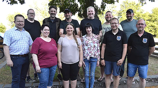 Elf Personen stehen für ein Gruppenfoto im Freien vor Bäumen und einem Zaun, wobei einige Freizeitkleidung und zwei schwarze Poloshirts mit Teublitzer Wappen tragen und gemeinsam in die Kamera blicken.