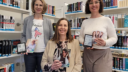 Drei Frauen stehen und sitzen zwischen Bücherregalen in einer Bibliothek und halten verschiedene Medien wie ein E-Book-Lesegerät, Flyer und Informationskarten in den Händen.