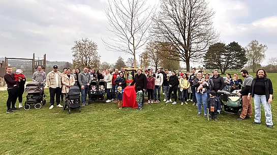 Eine große Gruppe von Familien mit Kindern und Kinderwagen versammelt sich auf einer Wiese rund um einen frisch gepflanzten Baum.